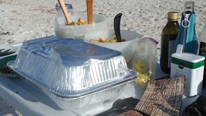 A picnic setup on a sandy beach with a covered food container, a cutting board with meat, and various food items and utensils. - Sun, beach, and great deals: Huge discounts on vacation destinations! (Cape Coral)