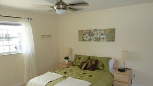 Une chambre à coucher avec un lit, des tables de chevet, un ventilateur de plafond, une fenêtre avec des rideaux et un tableau au mur.