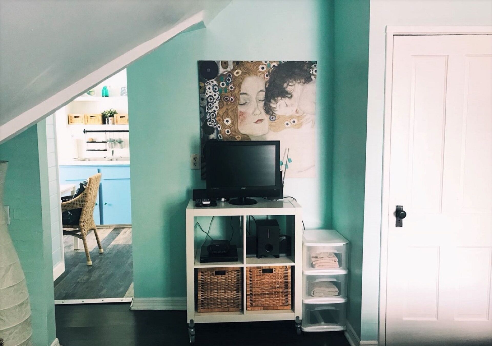 A room with teal walls, a white door, a television on a white stand, wicker baskets, and a wicker chair in the background.