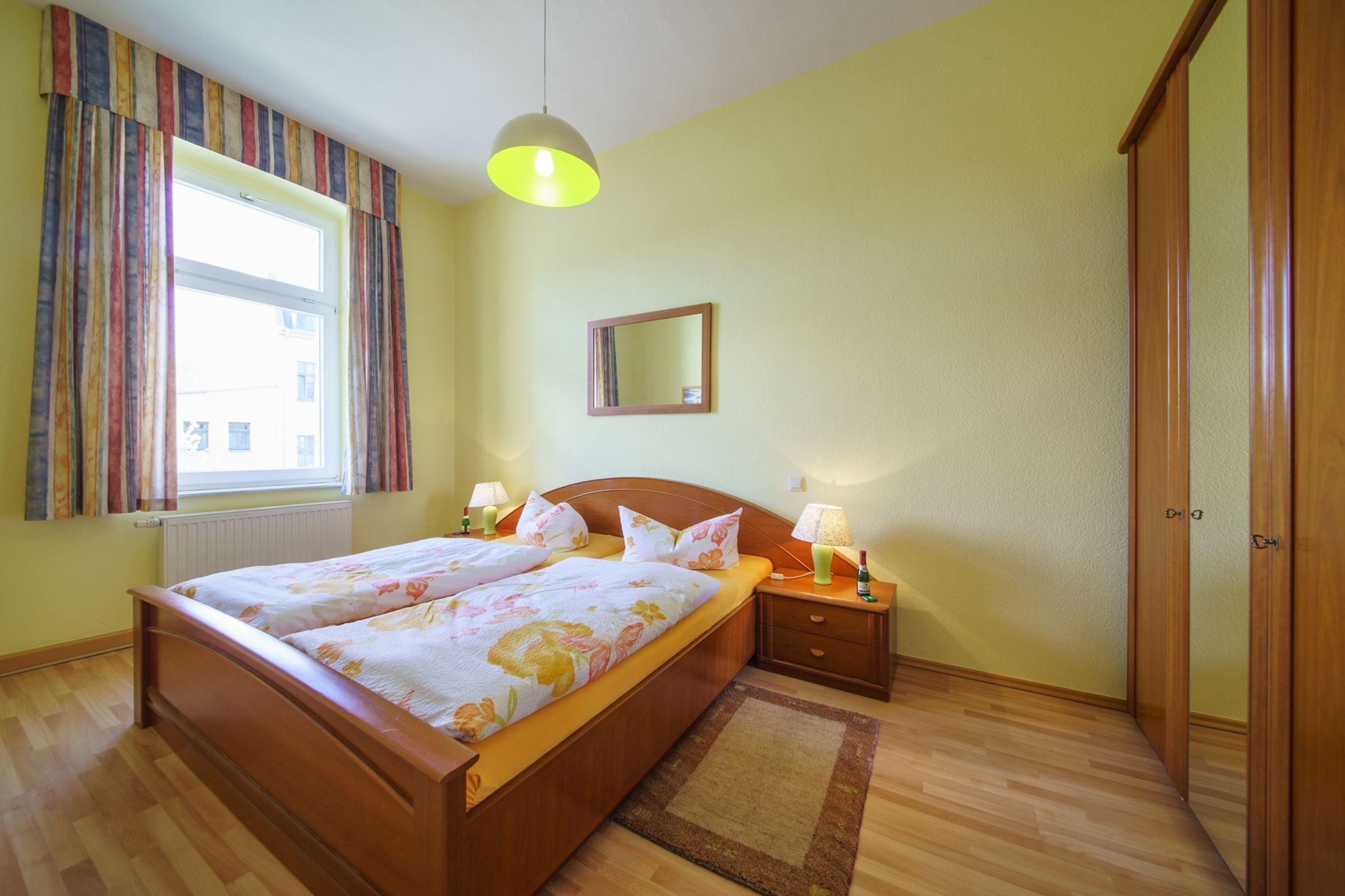 Een slaapkamer met een houten bed, twee nachtkastjes met lampen, een raam met gordijnen, een spiegel aan de muur en een kledingkast.
