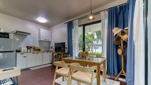 A modern kitchen with white cabinets, a dining table with chairs, and a large window with blue curtains. - Jasmine Garden Bungalow (Koh Samui)