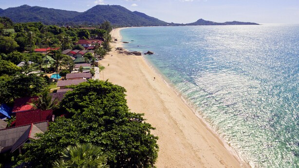 A beach with clear blue water, a sandy shore, and a backdrop of mountains. There are buildings with red roofs along the beach. - Jasmine Garden Bungalow (Koh Samui)