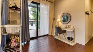 A room with a wooden floor, a white desk with a chair, a mirror, a hanging rack with clothes, and a view through a glass door. - Jasmine Garden Bungalow (Koh Samui)