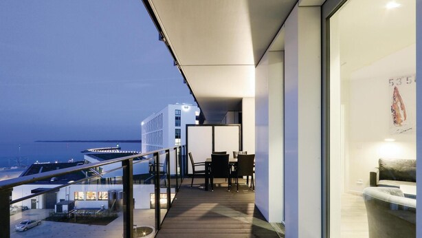 A balcony with a view of the sea, a dining area with tables and chairs, and a modern building in the background.
