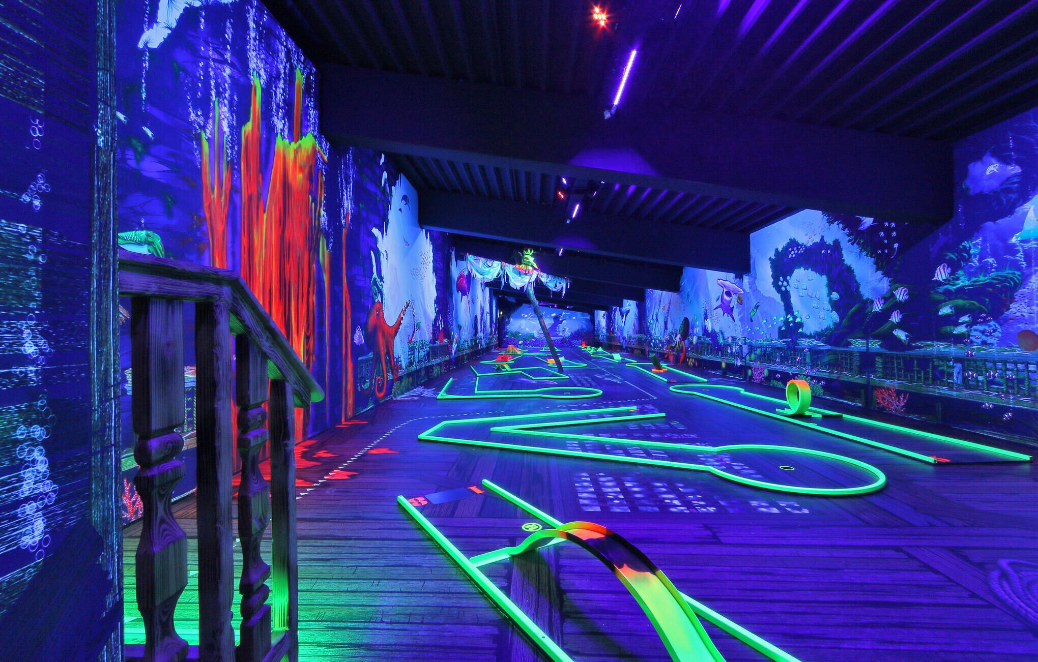 A neon-lit pathway with digital displays on the walls, a wooden deck, and a ceiling with lighting fixtures.