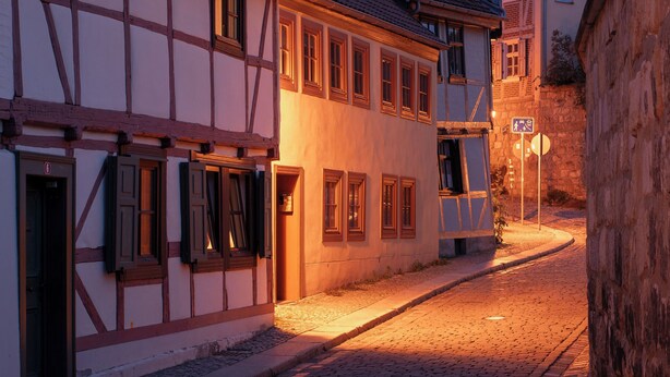 A narrow cobblestone street lined with traditional half-timbered buildings, featuring a mix of ground and first-floor windows, and a visible pedestrian crossing sign.
