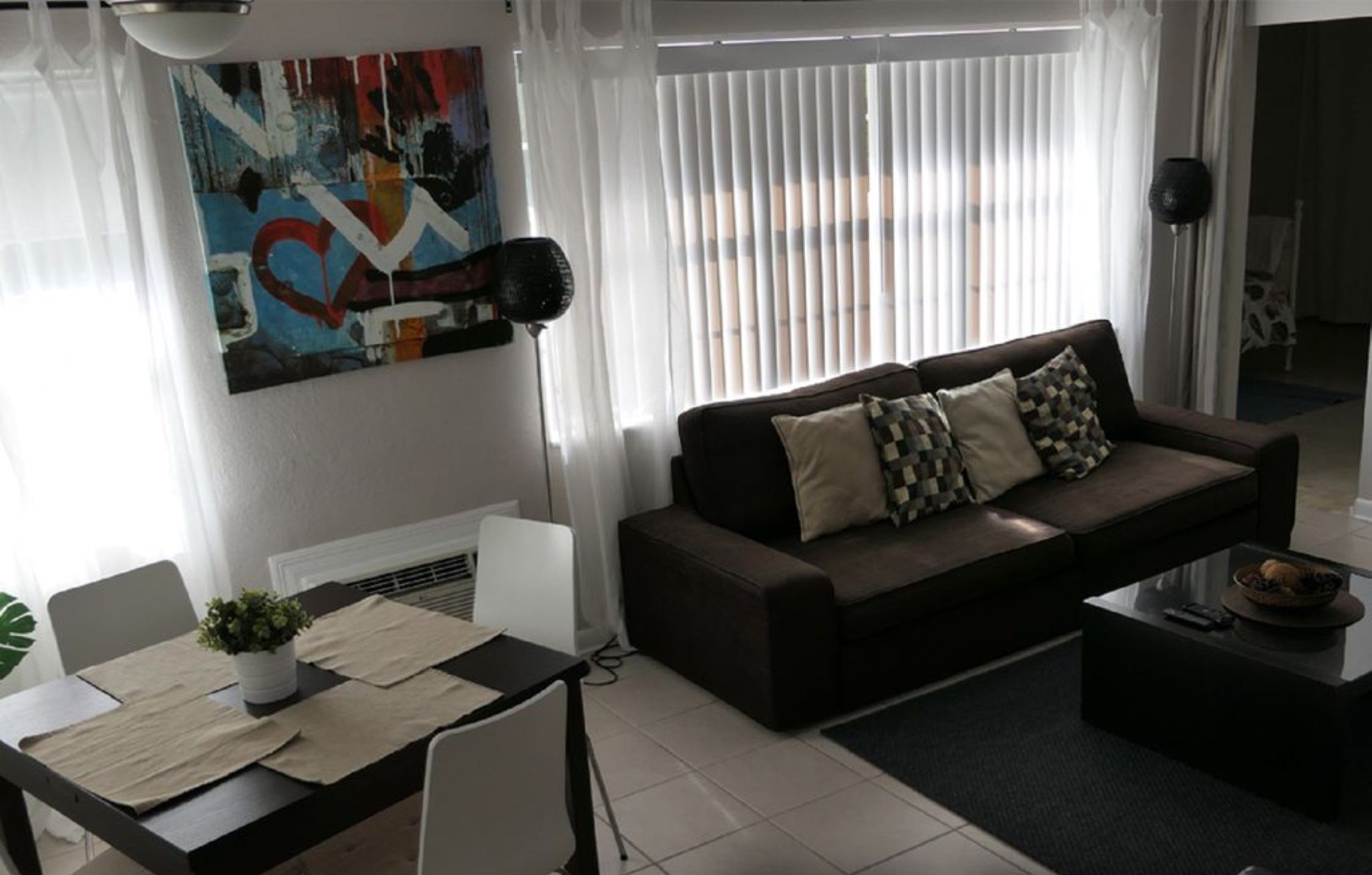 Een moderne woonkamer met een donkerbruine bank, witte en gemêleerde kussens, een eettafel met stoelen, en een groot abstract schilderij aan de muur.