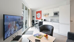 Un soggiorno moderno con una TV a schermo piatto, un'area pranzo con tavolo e sedie, una cucina con mobili bianchi ed elettrodomestici in acciaio inossidabile, e un'ampia finestra con tende.
