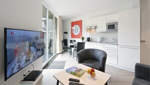 A modern living room with a flat-screen TV, a dining area with a table and chairs, a kitchen with white cabinets and stainless steel appliances, and a large window with curtains. - Dünenvilla 12, Whg 6 (Travemünde Waterfront)