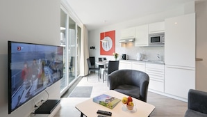 Ein modernes Wohnzimmer mit Flachbildfernseher, einem Essbereich mit Tisch und Stühlen, einer Küche mit weißen Schränken und Edelstahlgeräten sowie einem großen Fenster mit Vorhängen.