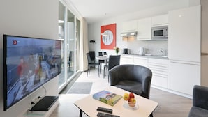 Een moderne woonkamer met een flatscreen-tv, een eethoek met tafel en stoelen, een keuken met witte kasten en roestvrijstalen apparatuur, en een groot raam met gordijnen.