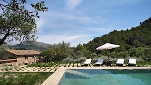 Ein Pool mit weißen Liegestühlen und einem Sonnenschirm, umgeben von viel Grün und einem Bergpanorama.