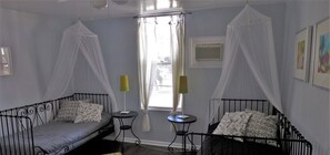Two single beds with patterned pillows are positioned opposite a small table in this cozy room, featuring a large window with white curtains and a patterned rug.