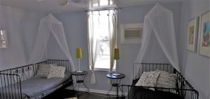 Two single beds with patterned pillows are positioned opposite a small table in this cozy room, featuring a large window with white curtains and a patterned rug.