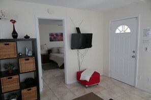 A room with a television mounted on the wall, a red chair, a white door, and a shelf with baskets and a vase.
