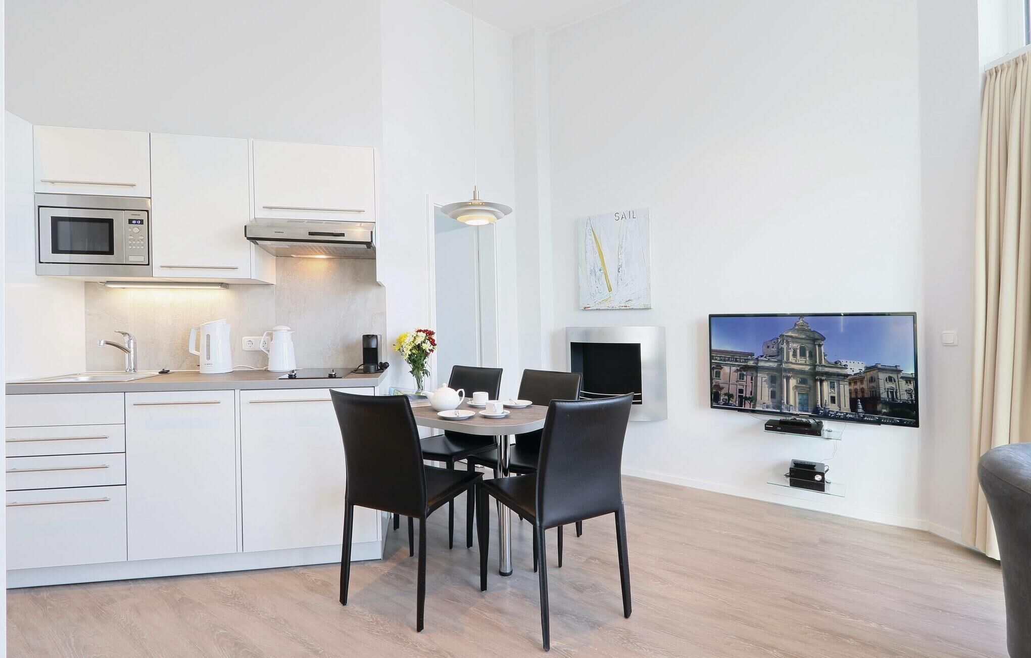 A modern kitchen with white cabinets, a microwave, and a coffee maker. There is a dining area with a table, chairs, and a vase of flowers. A painting of a building is on the wall, and a television is mounted above the table.