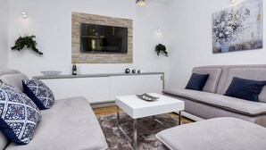 A modern living room with a grey sofa, a white coffee table, and a flat-screen TV mounted on a stone wall. There are two pendant lights, a painting on the wall, and a small plant on the windowsill. -  Holiday house with heated pool, terrace with sea view, 3 bedrooms, WiFi, BBQ (Bast)