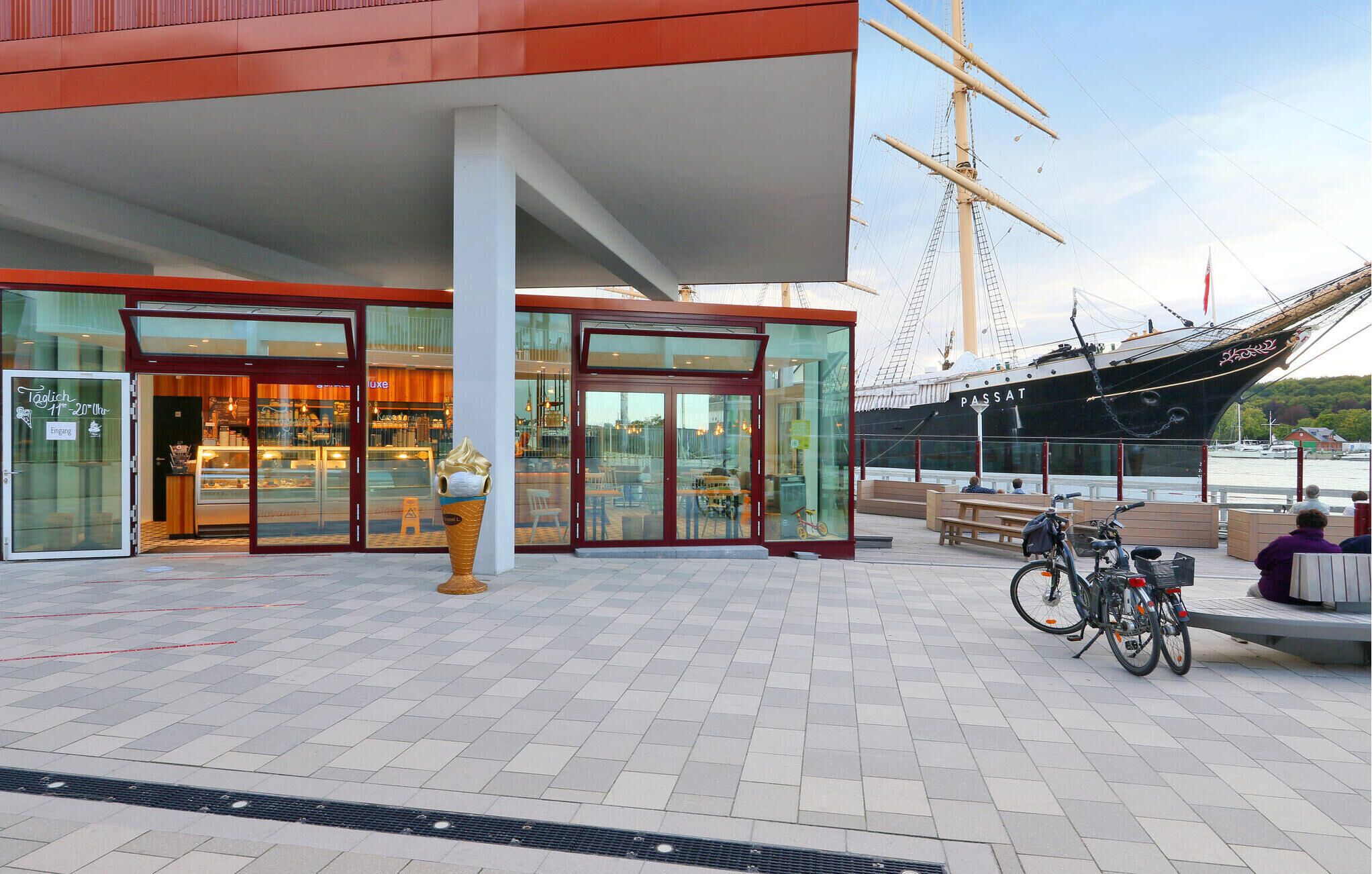 En modern byggnad med en fasad av glas, en stor skulptur i form av en glassvåffla utanför, och en segelbåt som ligger förtöjd i närheten.