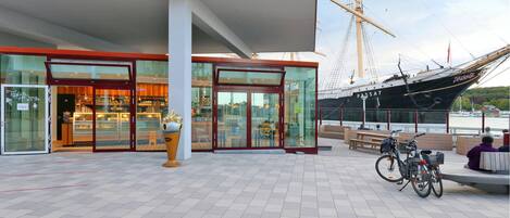 En modern byggnad med en fasad av glas, en stor skulptur i form av en glassvåffla utanför, och en segelbåt som ligger förtöjd i närheten.