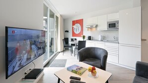 Un soggiorno moderno con una TV a schermo piatto, un'area pranzo con tavolo e sedie, una cucina con mobili bianchi ed elettrodomestici in acciaio inossidabile, e un'ampia finestra con tende.