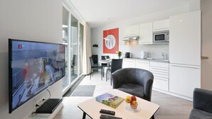 A modern living room with a flat-screen TV, a dining area with a table and chairs, a kitchen with white cabinets and stainless steel appliances, and a large window with curtains. - Dünenvilla 16, Whg 2 (Travemünde Waterfront)