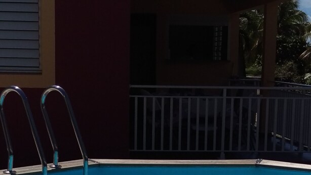 Una piscina con escaleras metálicas en un lateral, una zona cubierta con barandilla y un edificio de fachada roja al fondo.