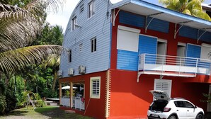 Una casa de dos pisos con fachada roja y azul, un coche blanco estacionado adelante y un pequeño jardín.