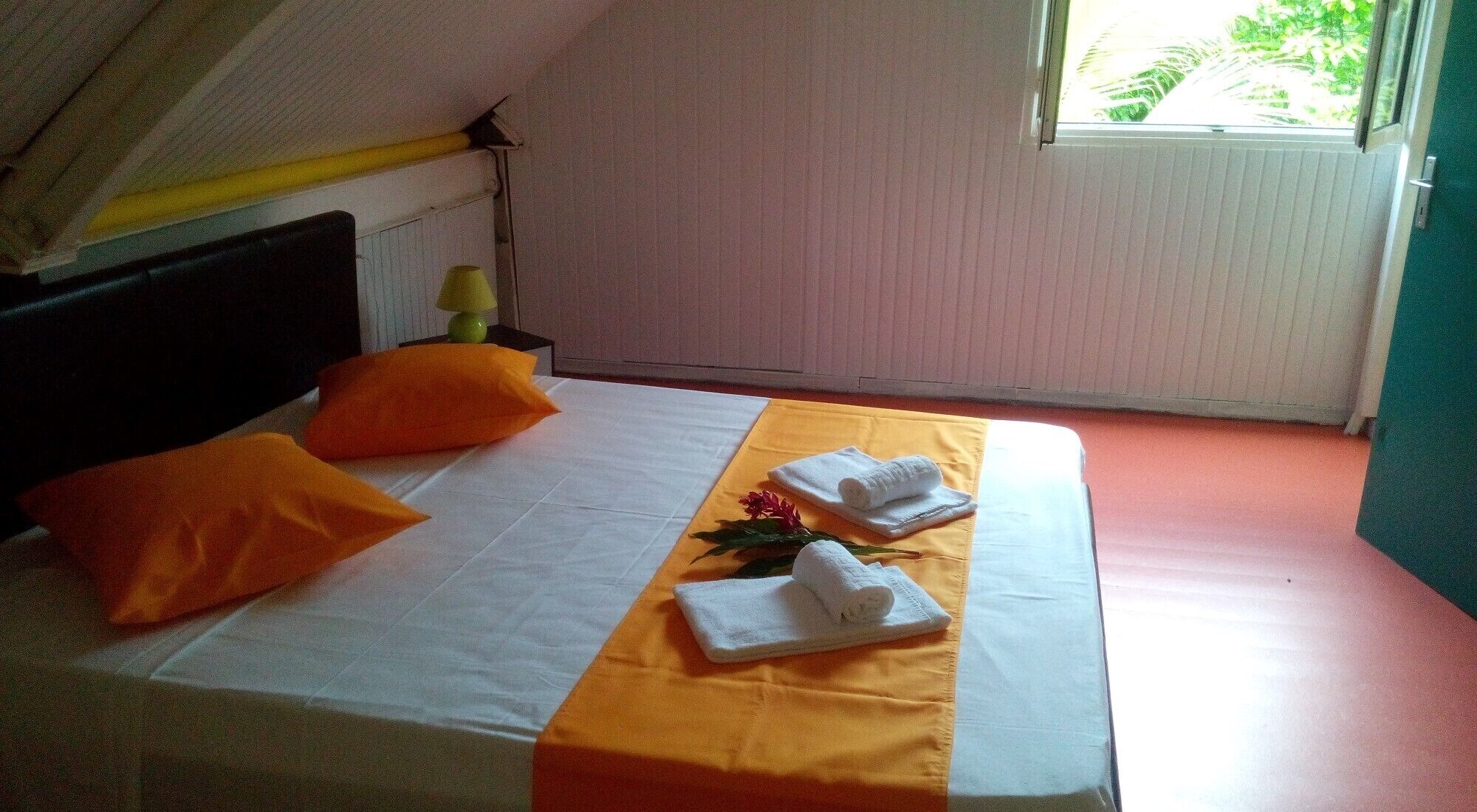 Un lit bien fait, agrémenté d’oreillers orange et d’une couvre-lit jaune, une petite table avec une lampe et une fenêtre donnant sur un paysage verdoyant.