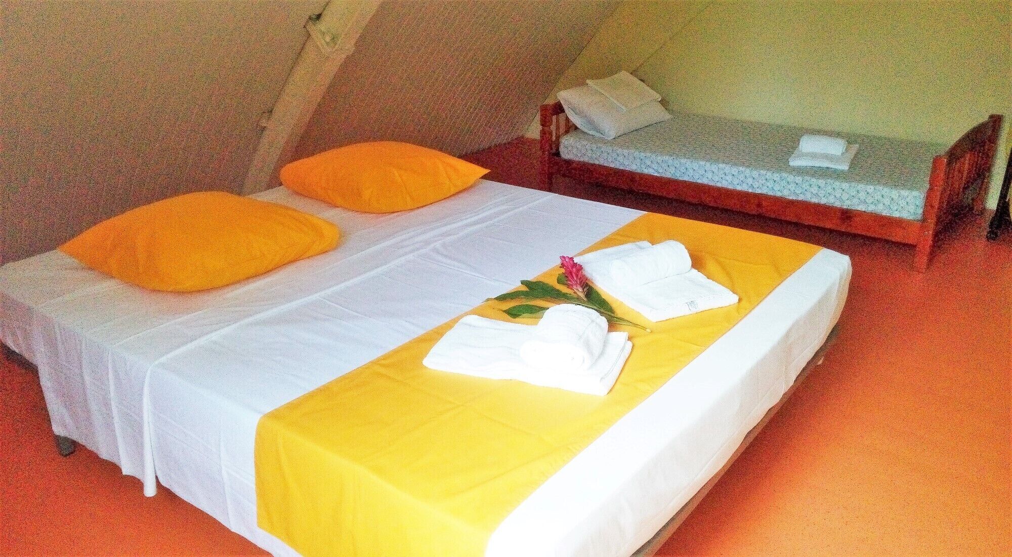 A single bed with a yellow bedspread and two orange pillows. There is a white towel and a pink flower on the bed.