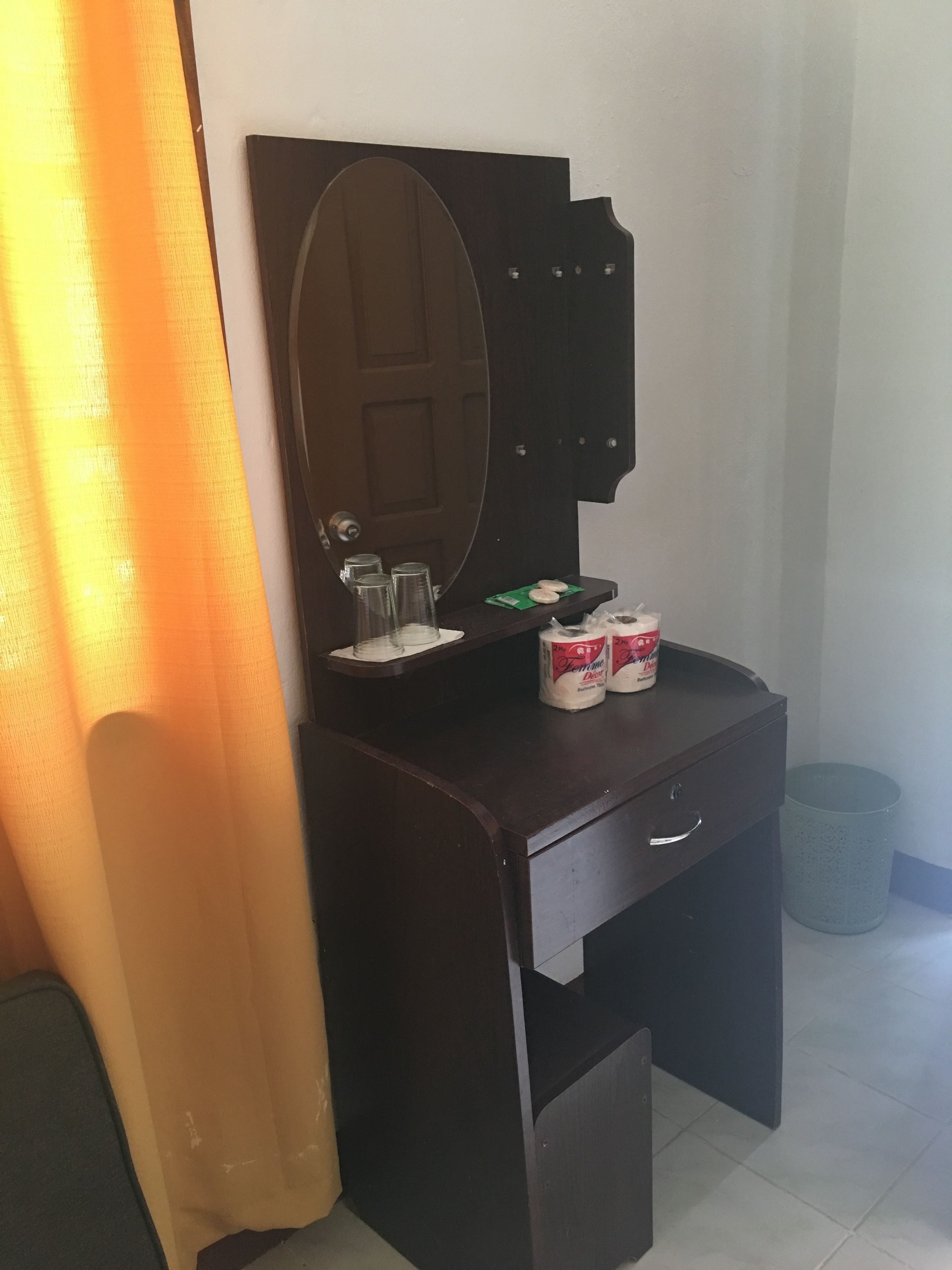 Uma escrivaninha de madeira escura com um espelho, dois rolos de papel higiênico e uma lixeira.