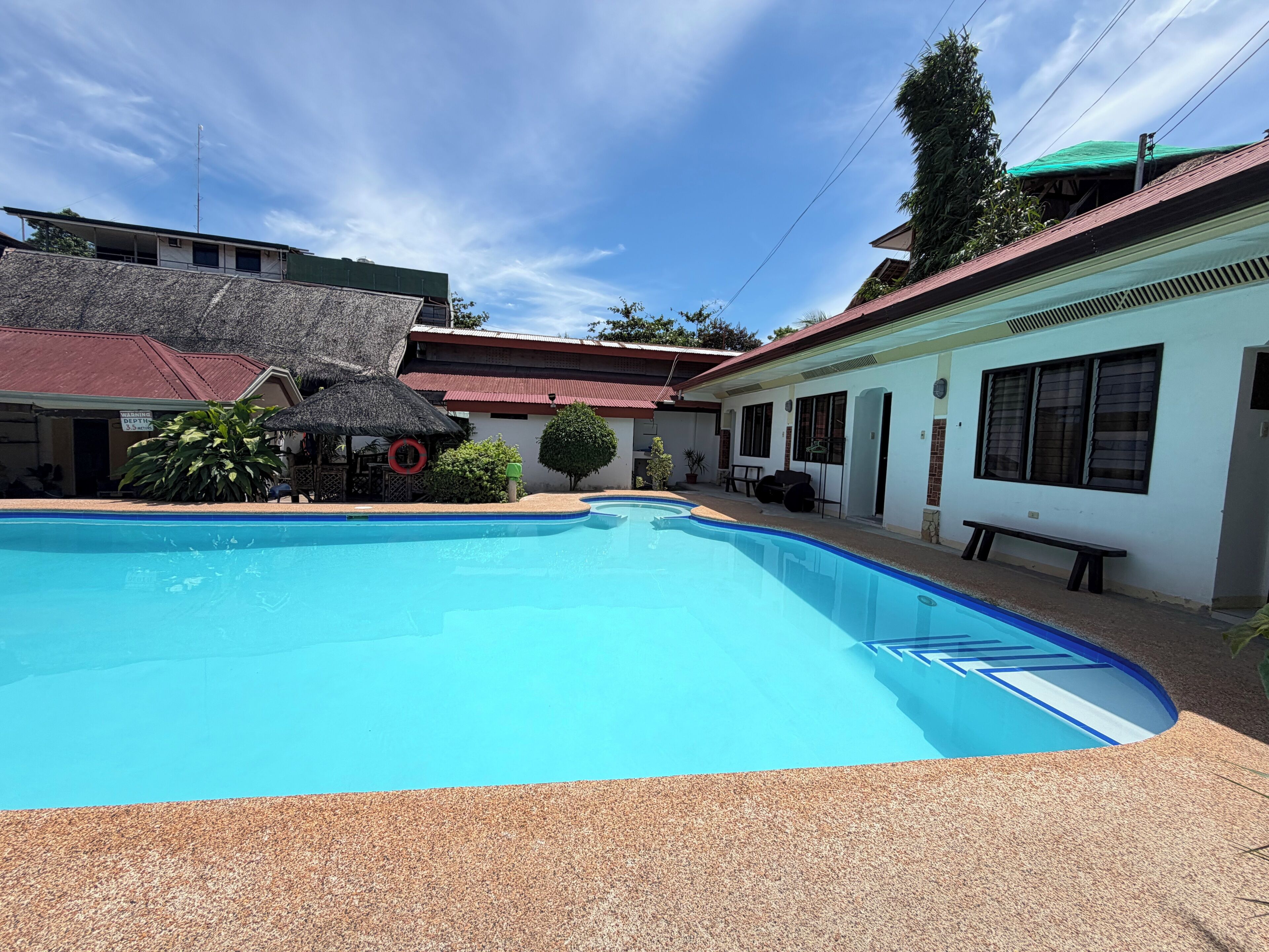 A swimming pool with a clear blue water surface, surrounded by buildings with red roofs and white walls.