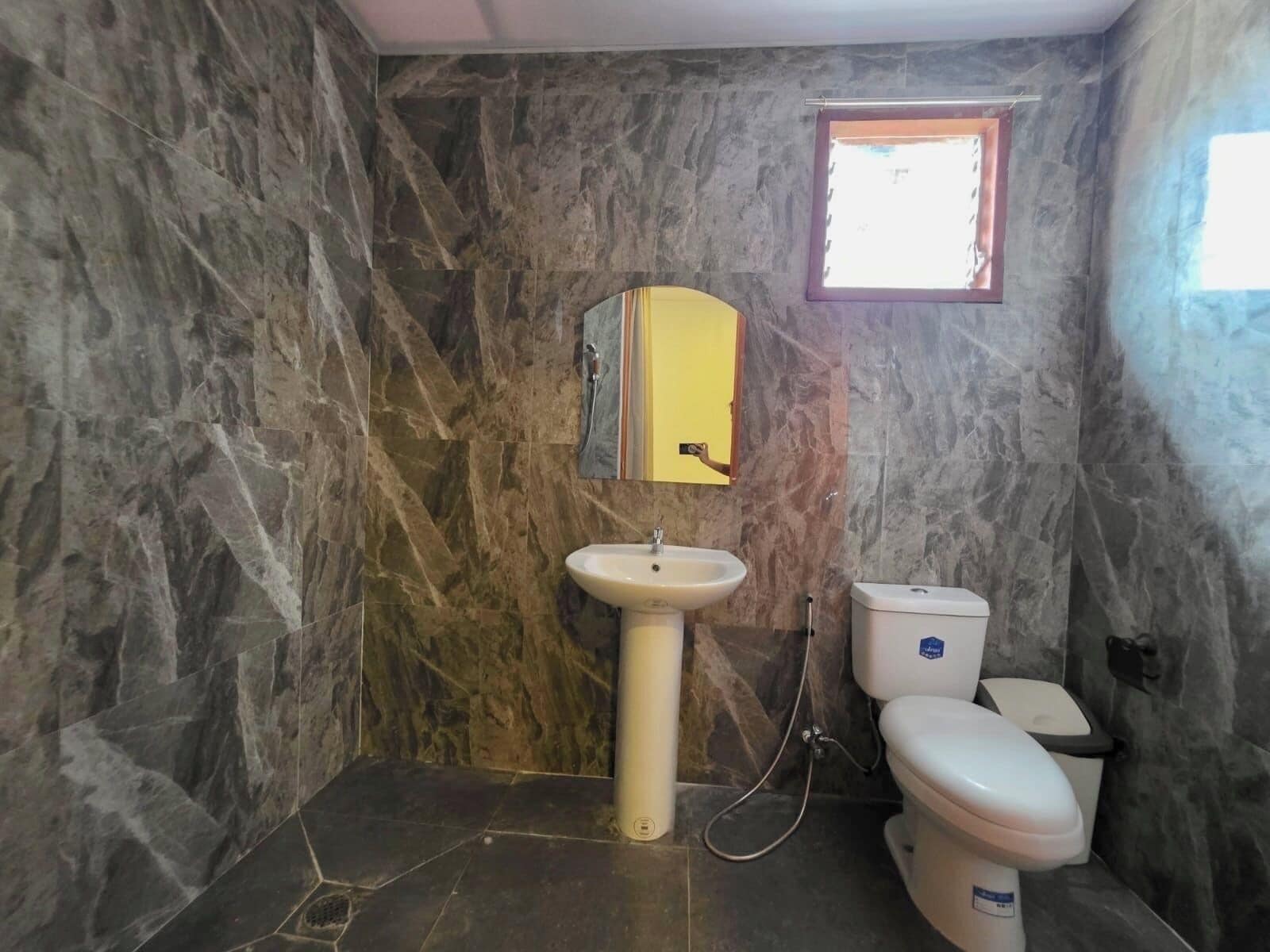 Une salle de bain avec un mur en marbre, un lavabo sur colonne, des toilettes et une fenêtre.