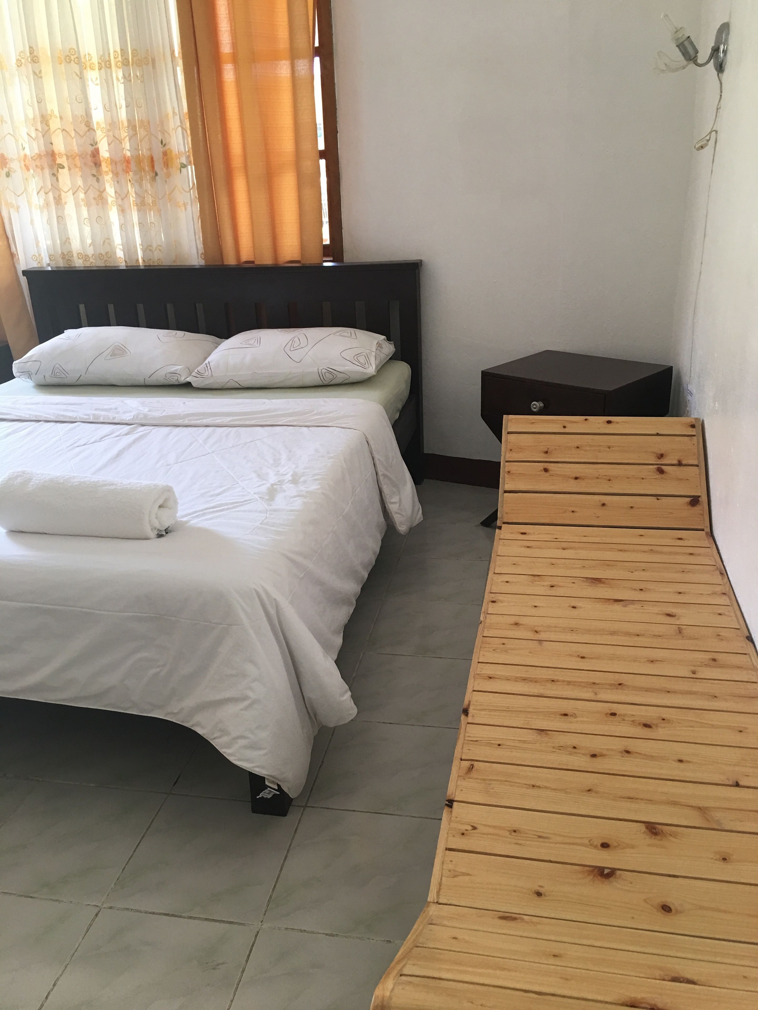 Een keurig opgemaakt bed met witte lakens en kussens, een houten bankje, een klein nachtkastje en een raam met gordijnen.