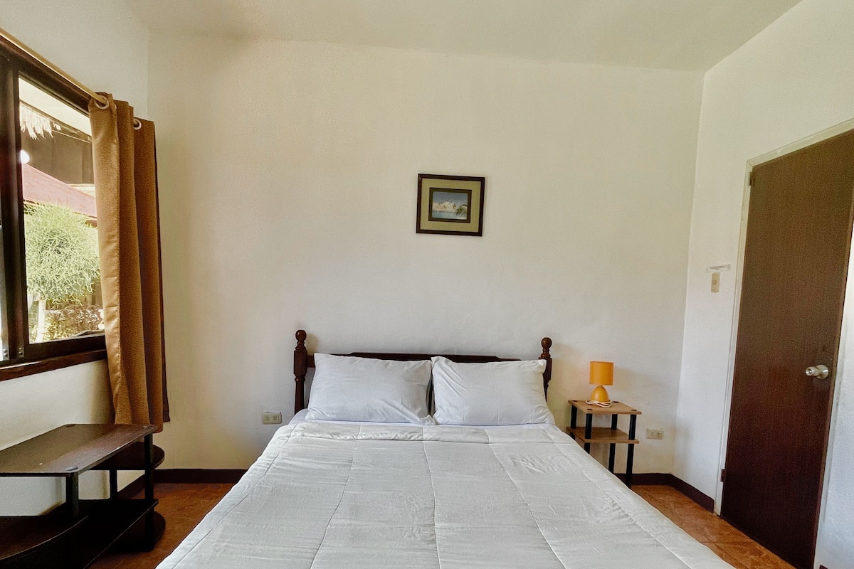 Un dormitorio con una cama grande, un banco de madera, una mesita con una lámpara y una puerta.