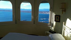 Hotelkamer met een bed, nachtkastje en drie gewelfde ramen met uitzicht op zee en een verafgelegen stad.