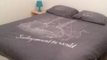 Un lit recouvert d’une couverture de couleur gris foncé ornée d’un motif représentant un voilier, et sur laquelle on peut lire le texte : « Naviguer autour du monde ».