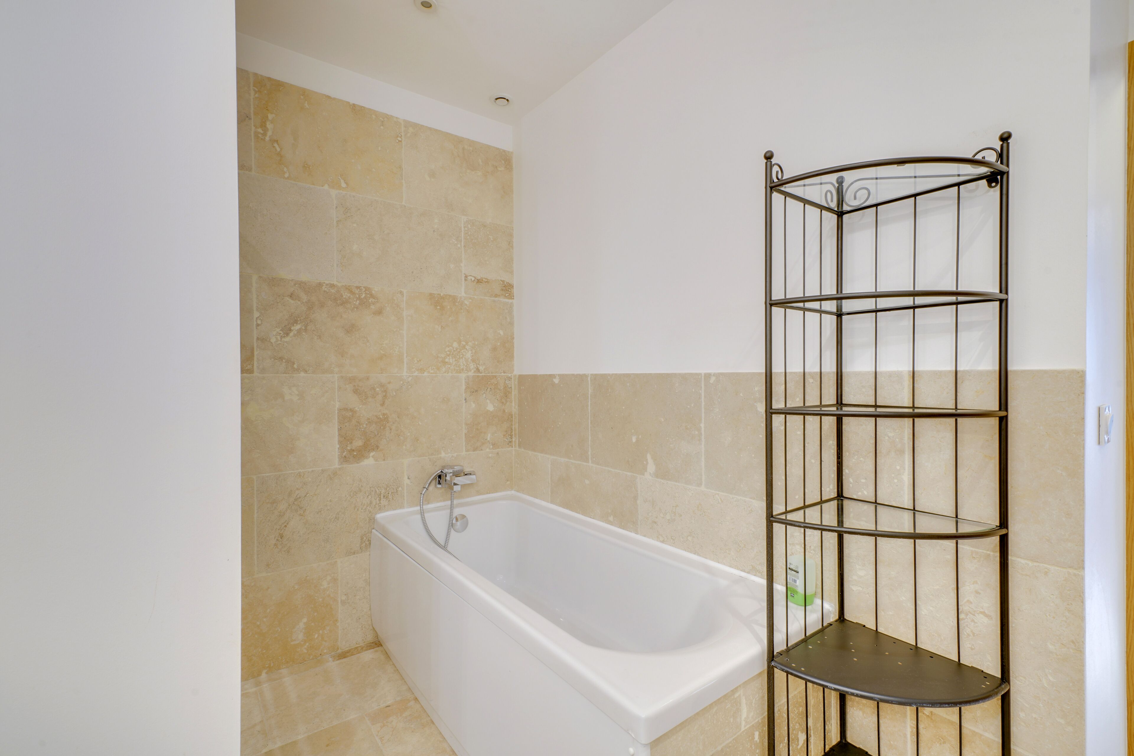 A bathroom with a white bathtub, beige tiled walls, and a metal shelf unit.