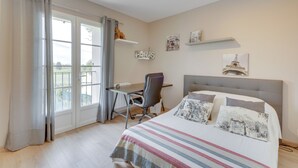 Une chambre à coucher avec un lit, un bureau et une chaise. Il y a deux fenêtres avec des rideaux, un luminaire au plafond et des œuvres d’art accrochées au mur.