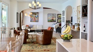 A spacious living room with a sofa, armchairs, a coffee table, and a chandelier. There is a dining area with chairs and a table, and a kitchen with a fruit bowl and bananas. - Luxury  Villa with heated pool, spa/hot tub, dock, near beach,  Sunsets! (St. Augustine)