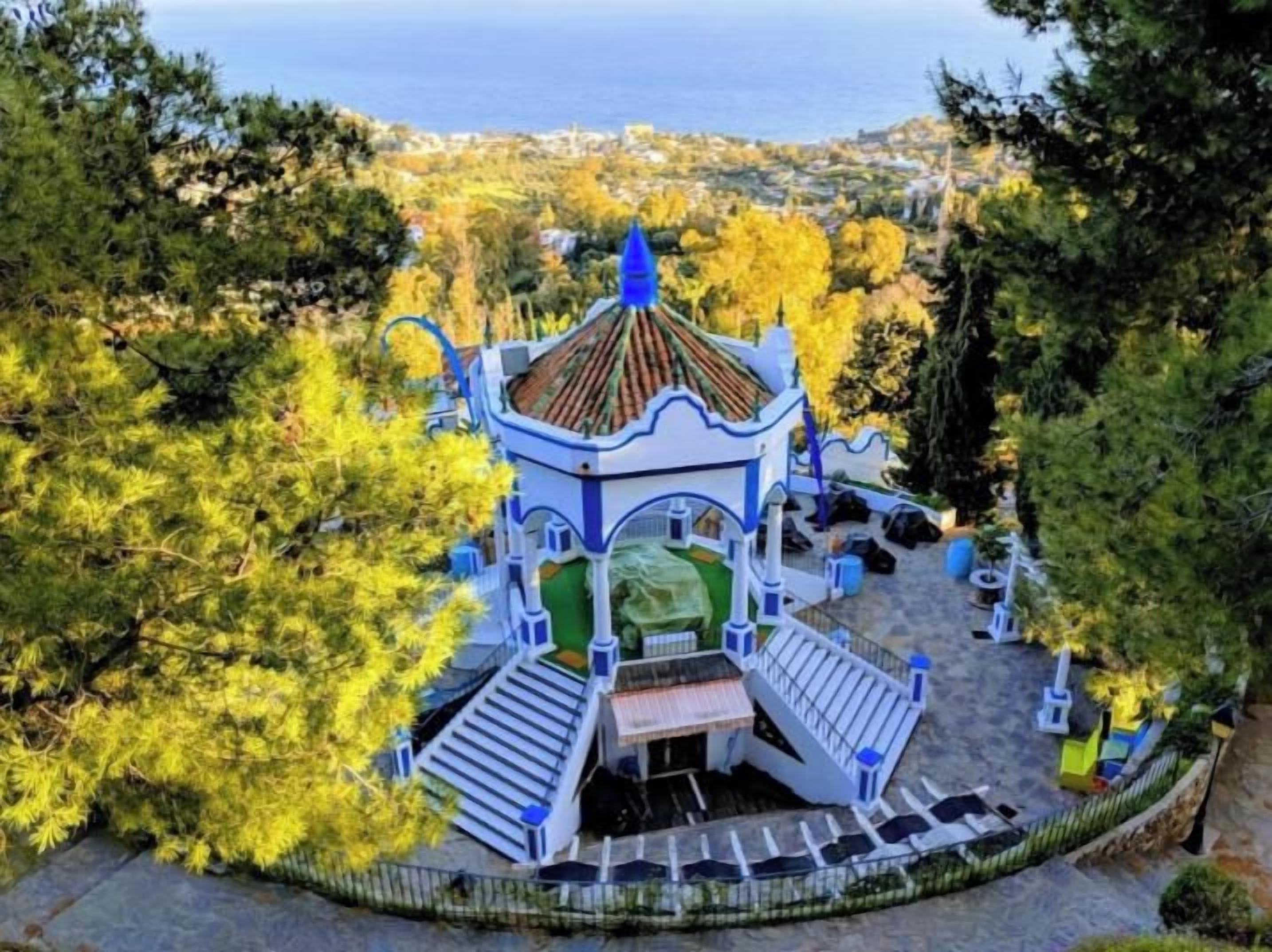 A gazebo-style structure with a tiled roof, surrounded by trees and a view of the sea.
