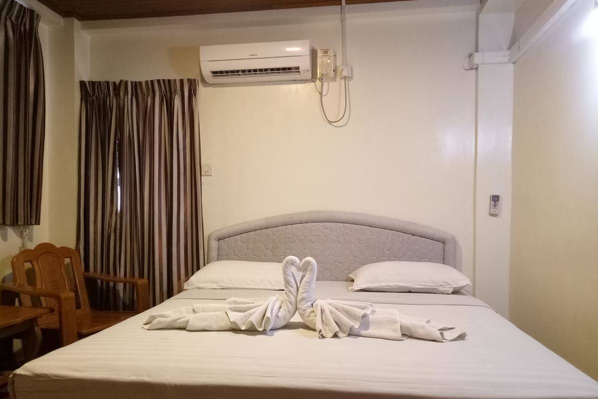 A hotel room with a bed, a wooden chair, a window with curtains, and an air conditioning unit.
