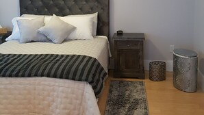 A bedroom with a tufted headboard, a bed with striped bedding, a bedside table with a lamp, a waste bin, and a patterned rug. - Right in the Quaint Village of Hammondsport (Hammondsport)