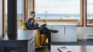A modern interior with a person sitting by a large window overlooking a beach. - The SkyHouse- Life as Art- Sauna Available (Grand Marais)