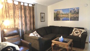 Una sala de estar con un sillón de cuero, un sofá, una mesa de centro y un tapete estampado. Hay dos cuadros colgados en la pared y una fotografía enmarcada.