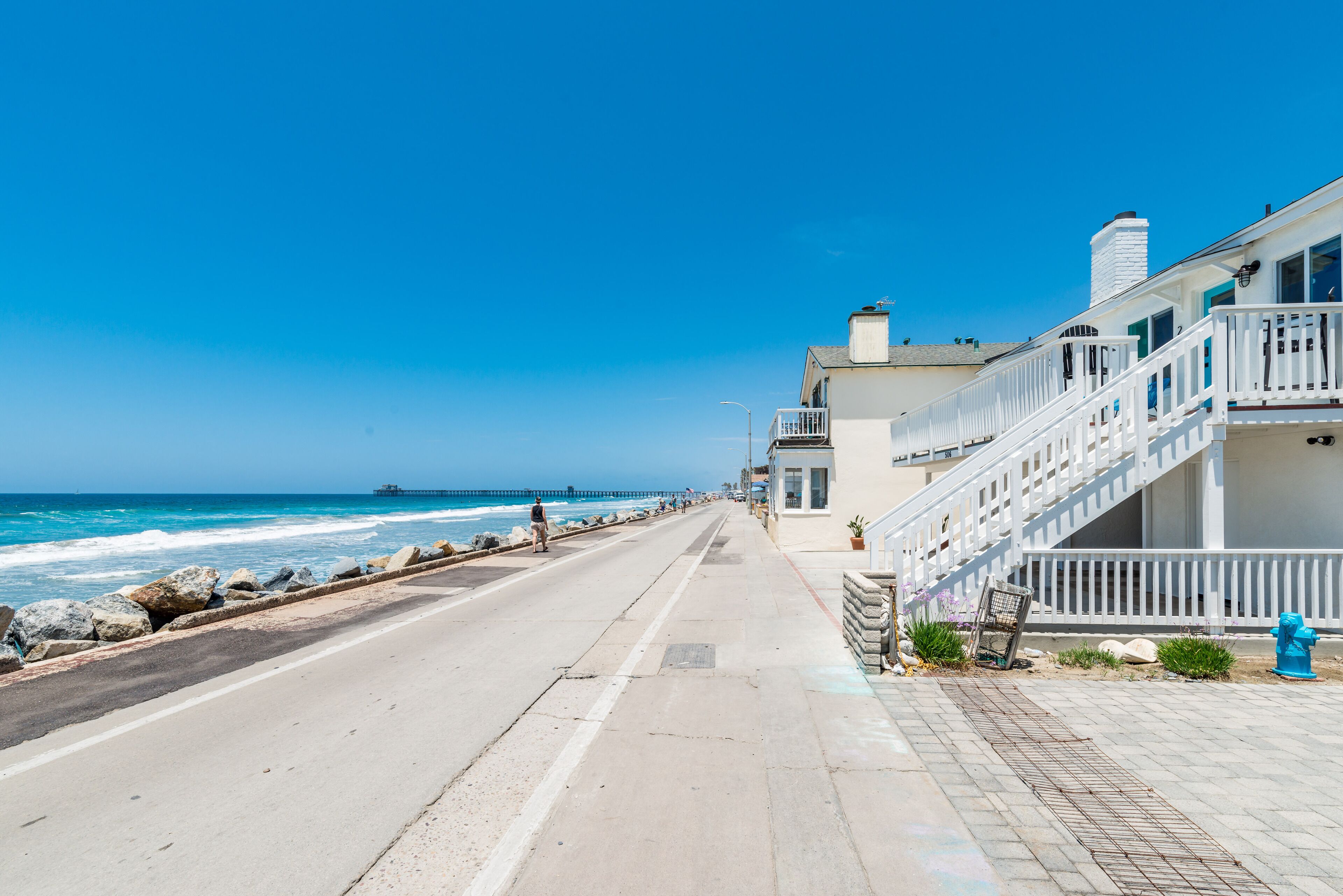 Une route côtière avec un trottoir, une borne d’incendie bleue et un bâtiment doté d’une balustrade et d’un escalier blancs.