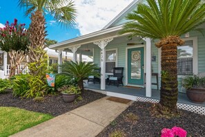 A two-story house with a porch, a front door, and a small garden area with plants and a palm tree.