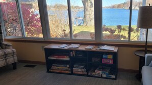 A room with a large window overlooking a lake, a bookshelf with board games, a plaid chair, and a lamp.