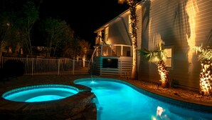 Una piscina con diseño curvo, rodeada por una valla blanca, y una casa con balcón y televisor montado en la pared.