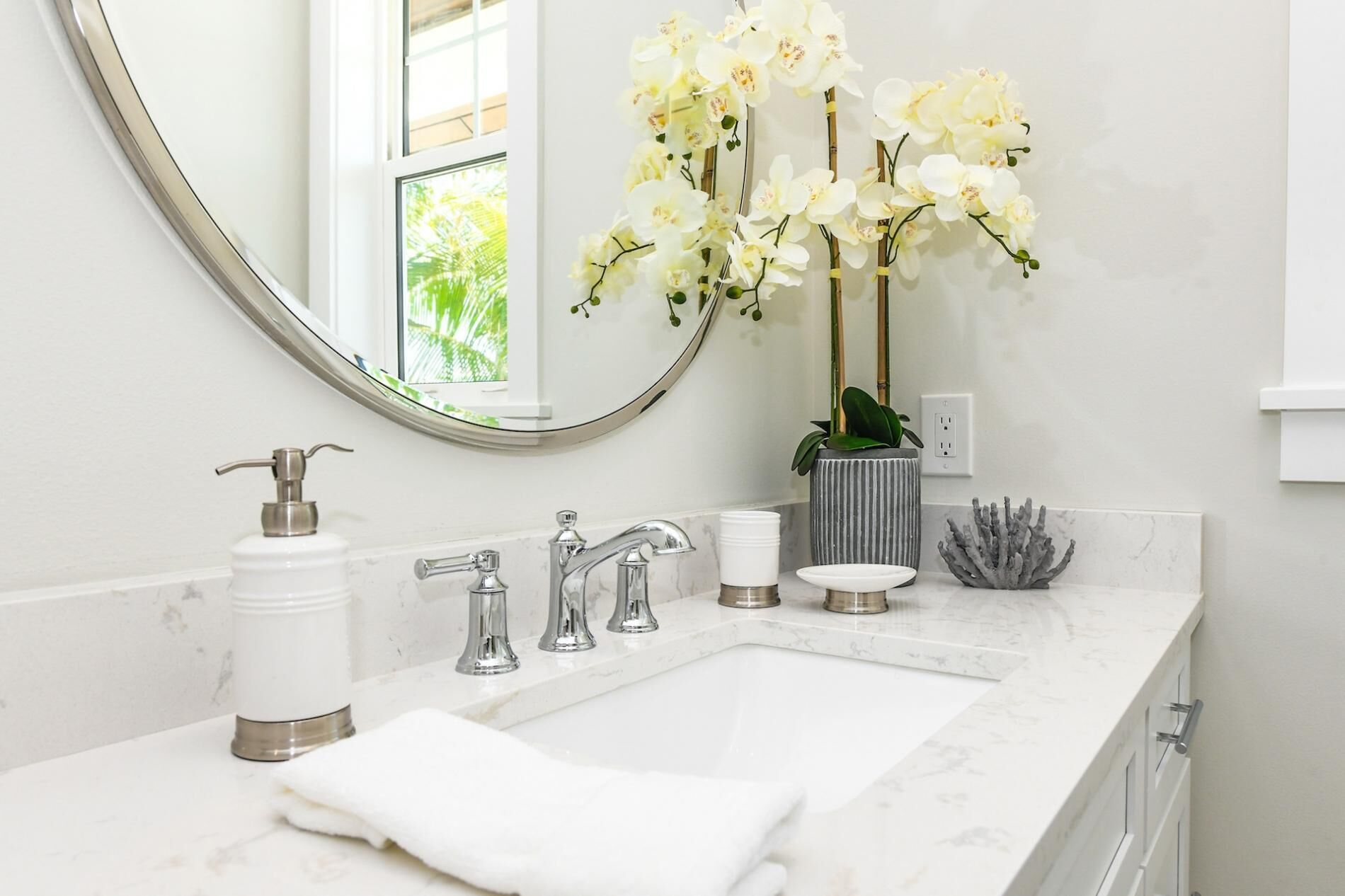A bathroom with a white marble sink, a large mirror, a potted orchid, and a coral decoration.