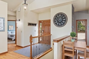Un escalier avec une rampe en bois mène à un palier où se trouvent une table à manger et des chaises. Une grande horloge murale est fixée au-dessus de la table.
